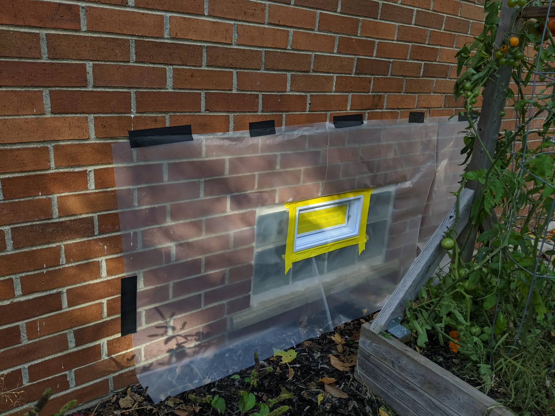 Large clear plastic mask taped over a brick wall around a small white glass-block window vent.