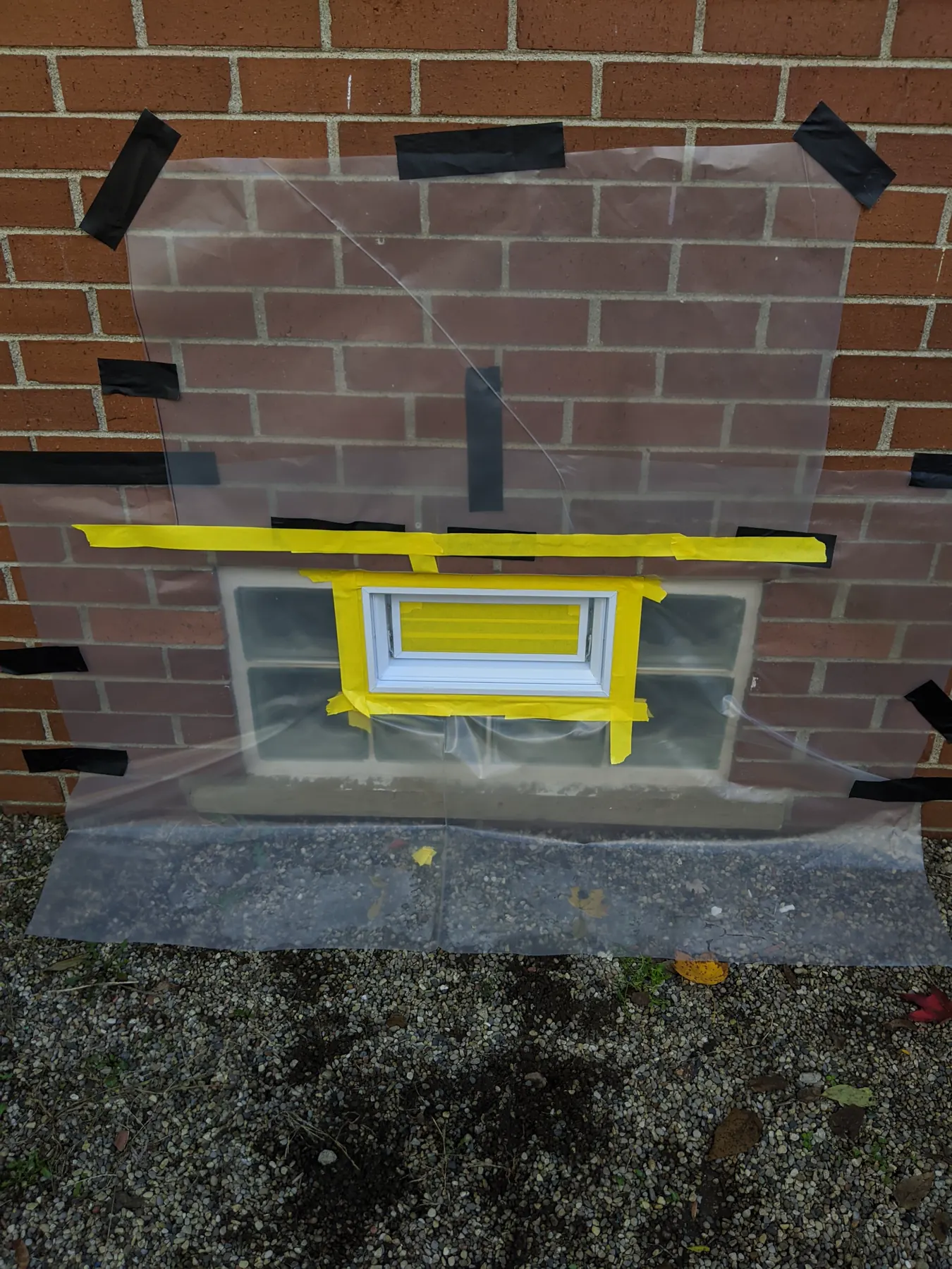 Painter's tape closely masking a white vent within a larger plastic-covered glass-block opening.