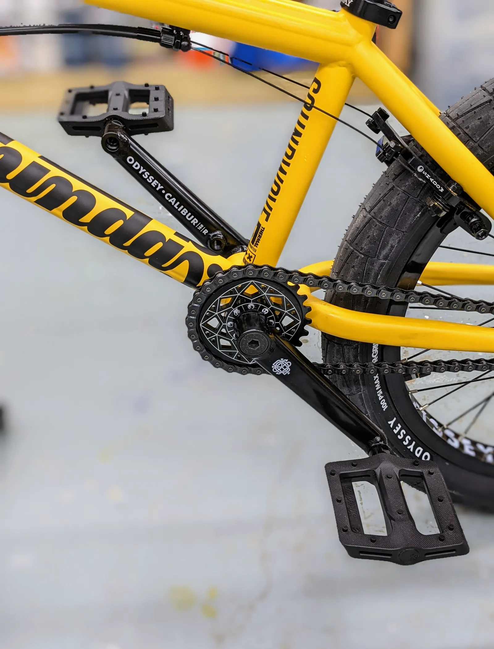 Close view of the installed black sprocket, crank, pedal, chain, and yellow frame.
