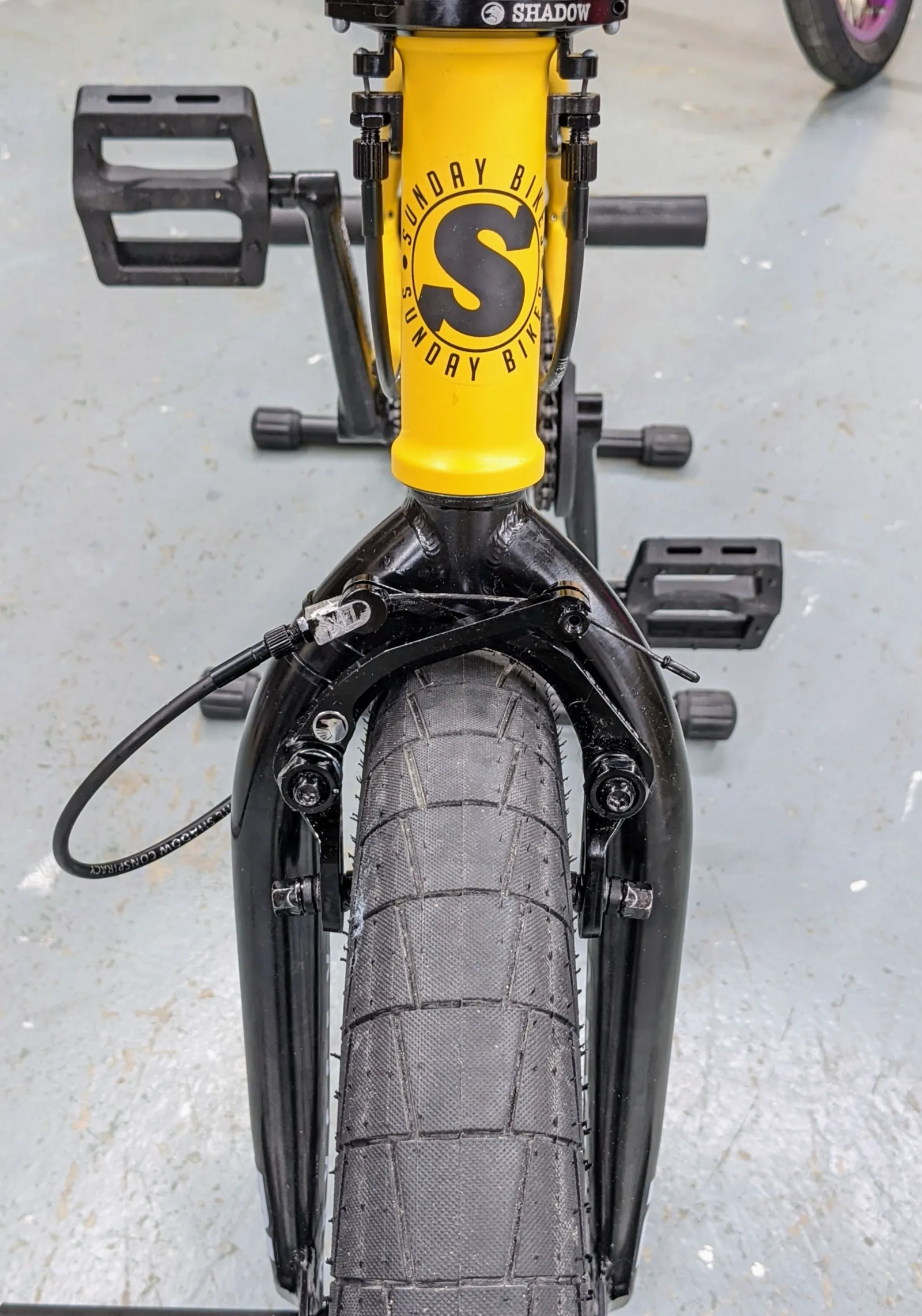 Centered close view of the yellow Sunday headtube, black stem, pedals, fork, pegs, and front tire.