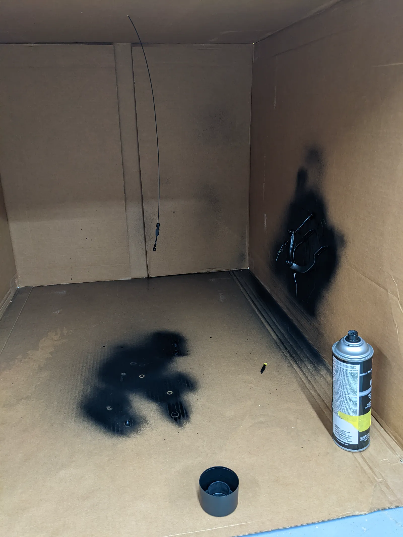 Black overspray around a small part inside a cardboard spray booth, with an aerosol can nearby.