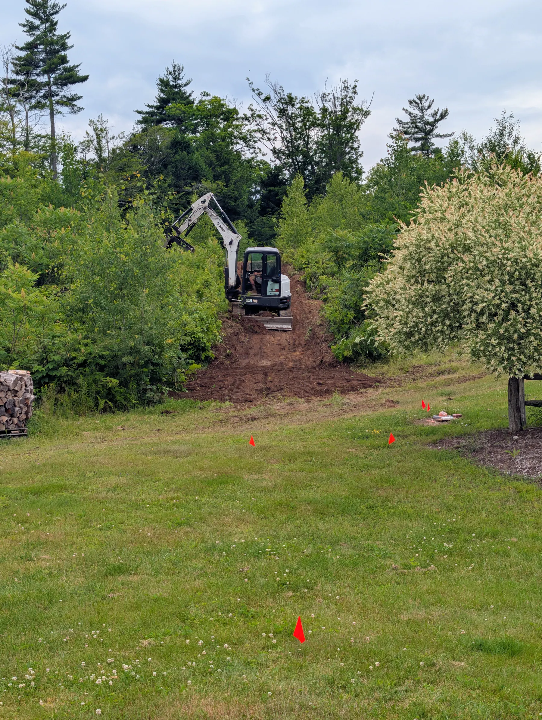 Excavator cutting the straight slide path down the hill toward orange survey flags in the backyard.