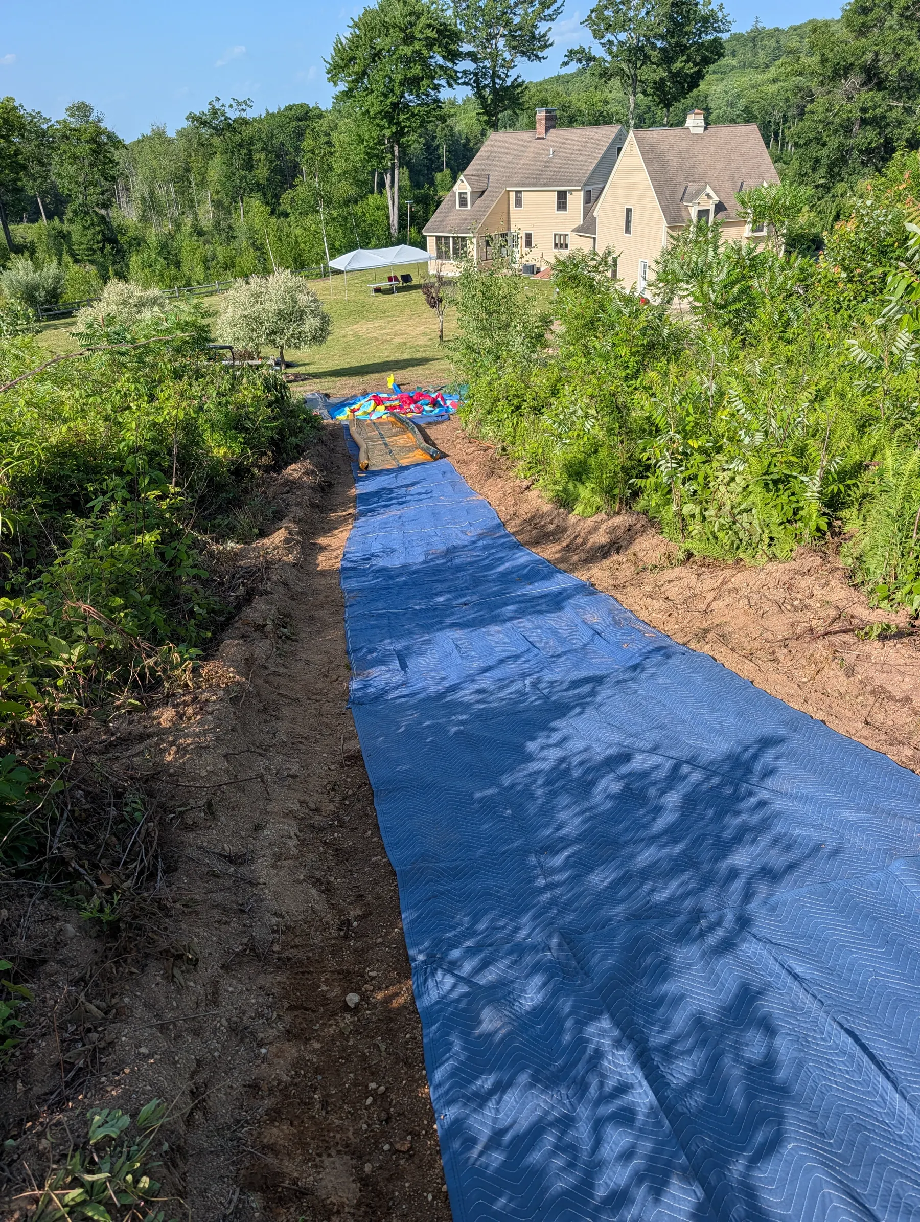 Blue moving blankets laid continuously down the excavated slide corridor toward the bounce castle.