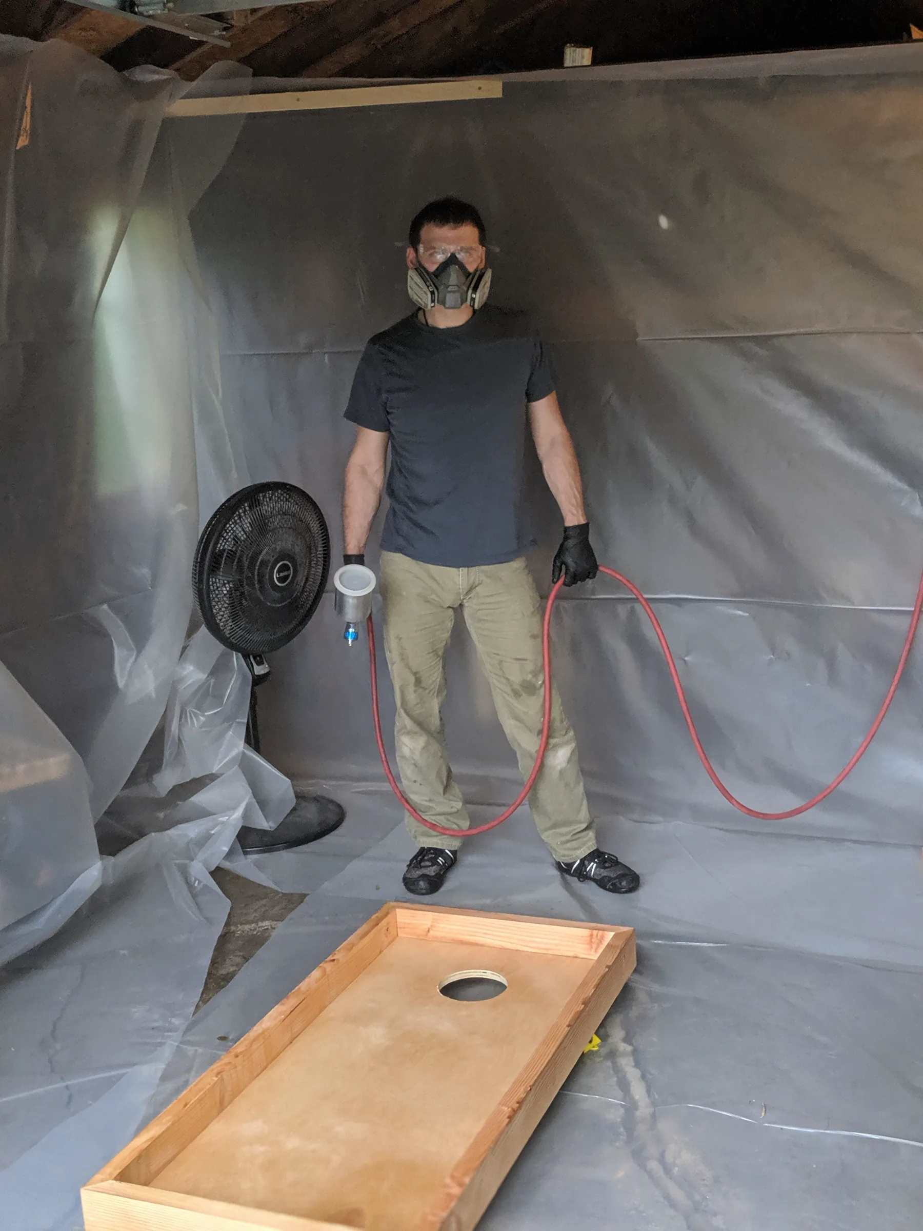 Builder wearing a respirator and holding spray equipment beside a cornhole board in the plastic-lined finish booth.