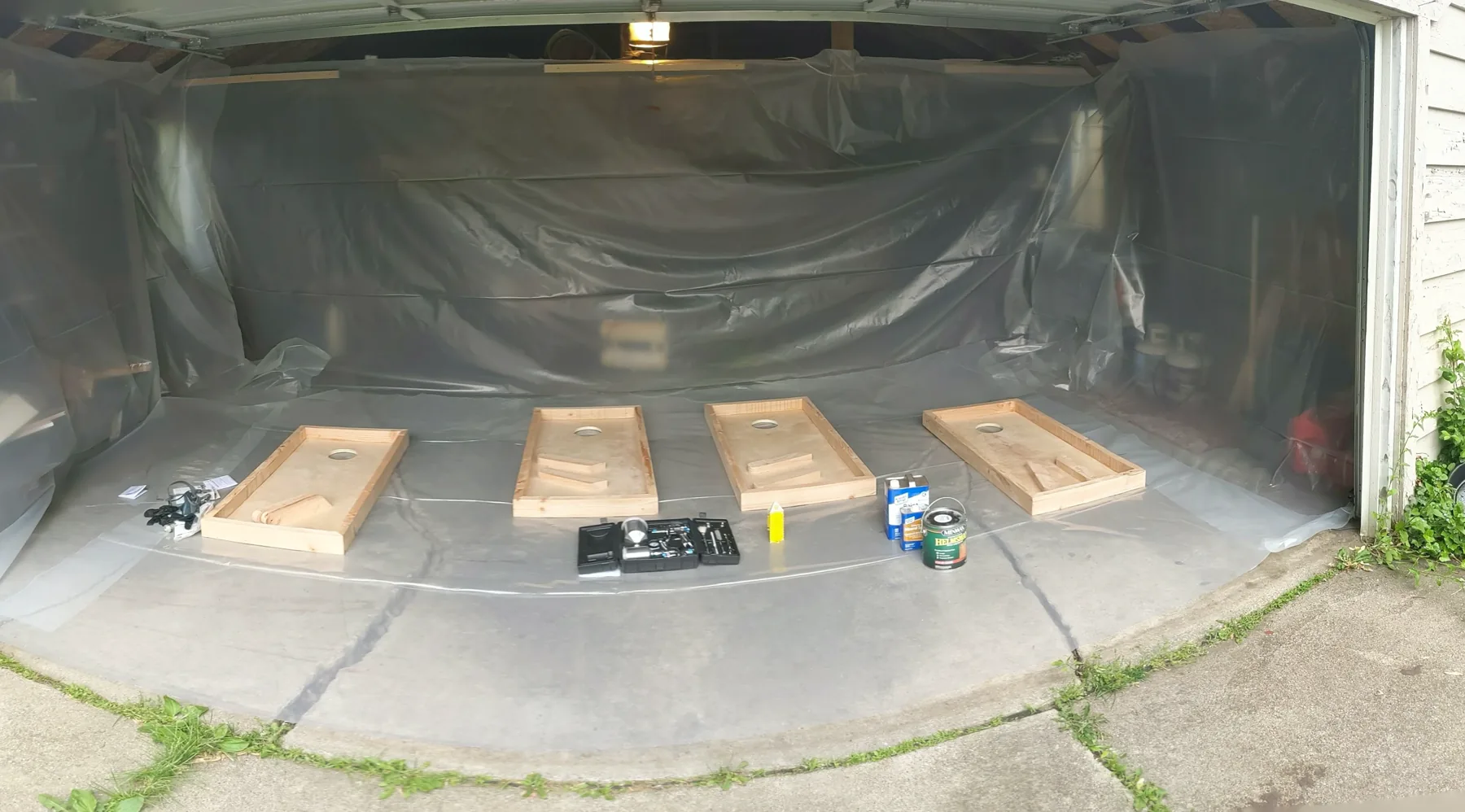 Panoramic view of four cornhole boards arranged in a plastic-lined garage finish booth with spray equipment between them.