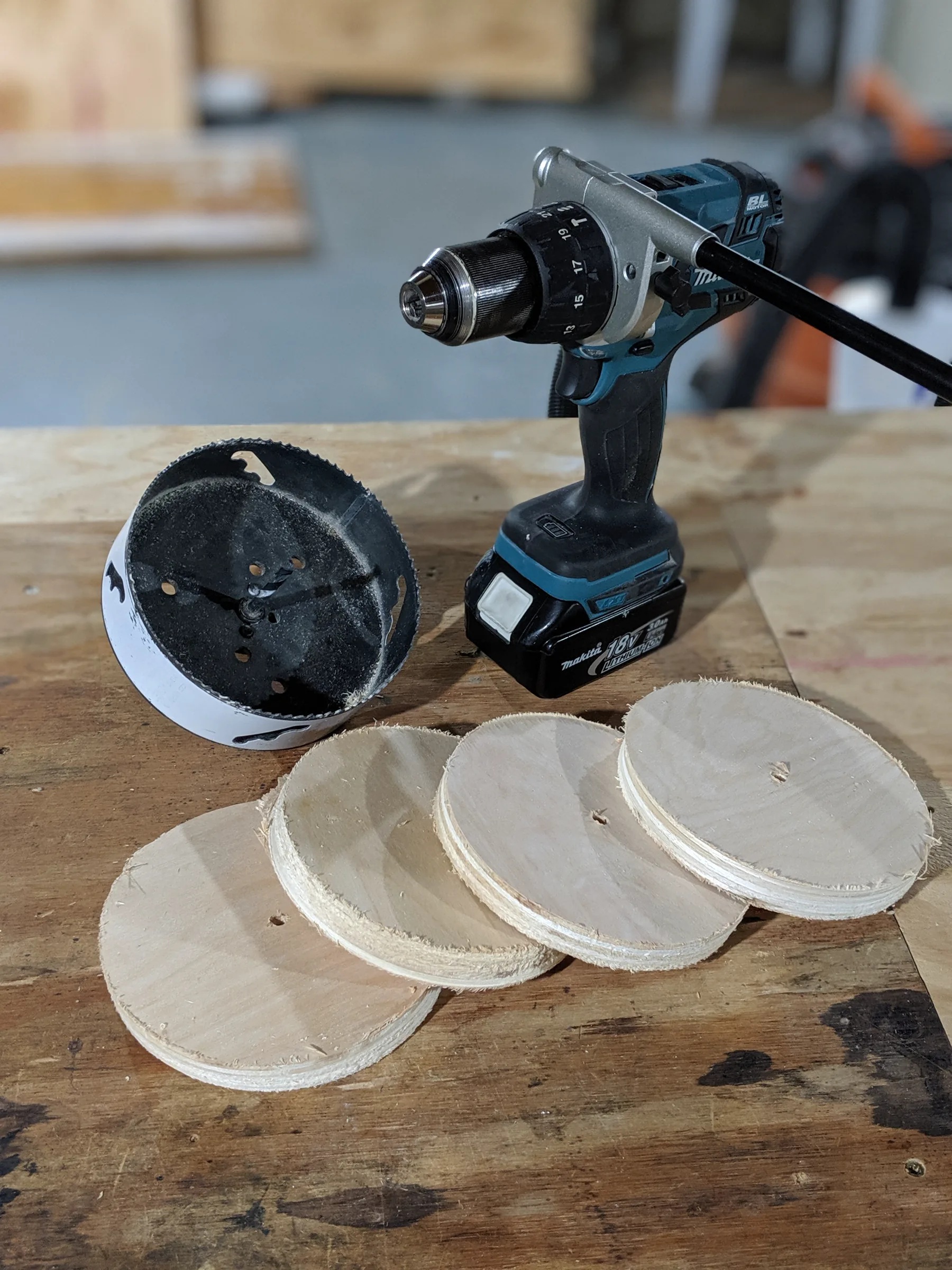 Drill, hole-cutting tool, and round birch cutouts on a workbench.