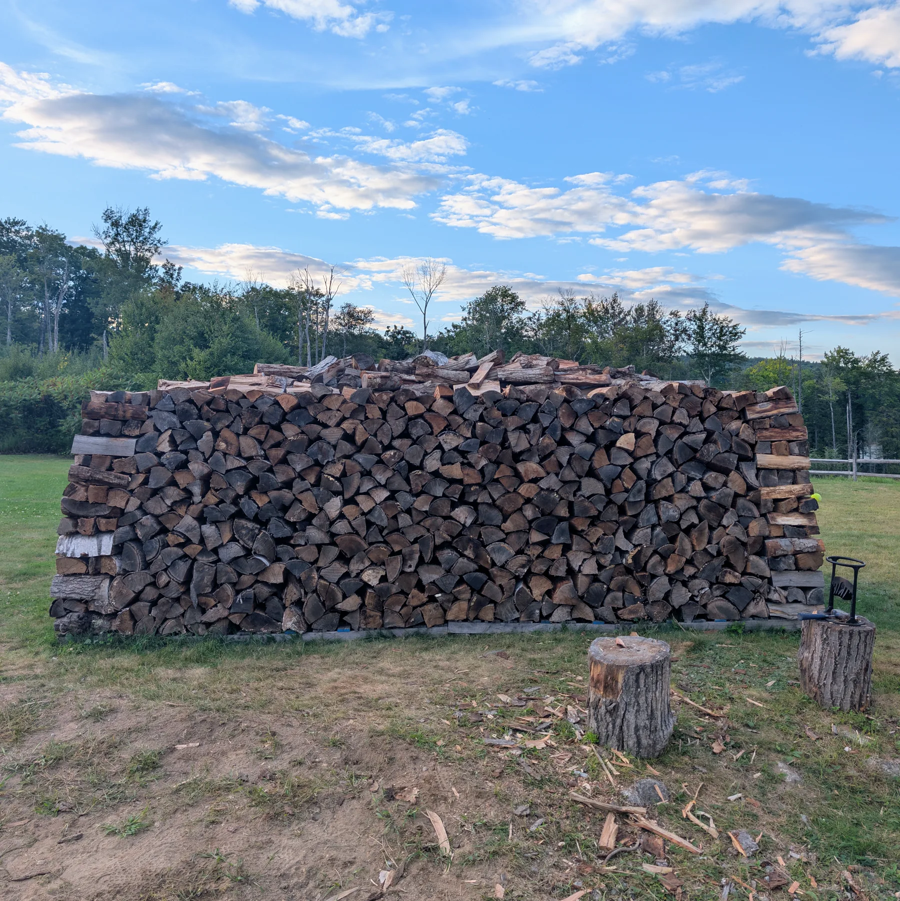 Completed rectangular firewood stack with a fully closed long wall, inward-tapered corner towers, and a shallow crown along the top.