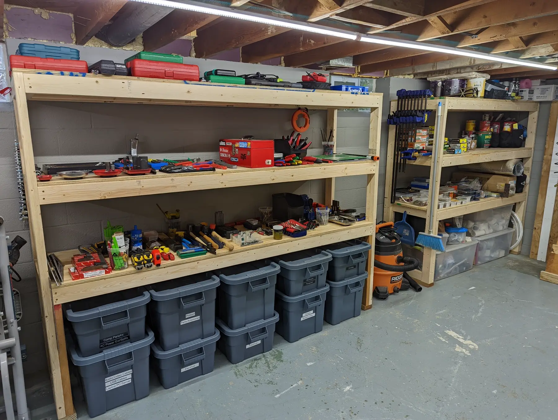 Completed long basement workshop shelving fully stocked with tools, cases, and labeled storage bins.